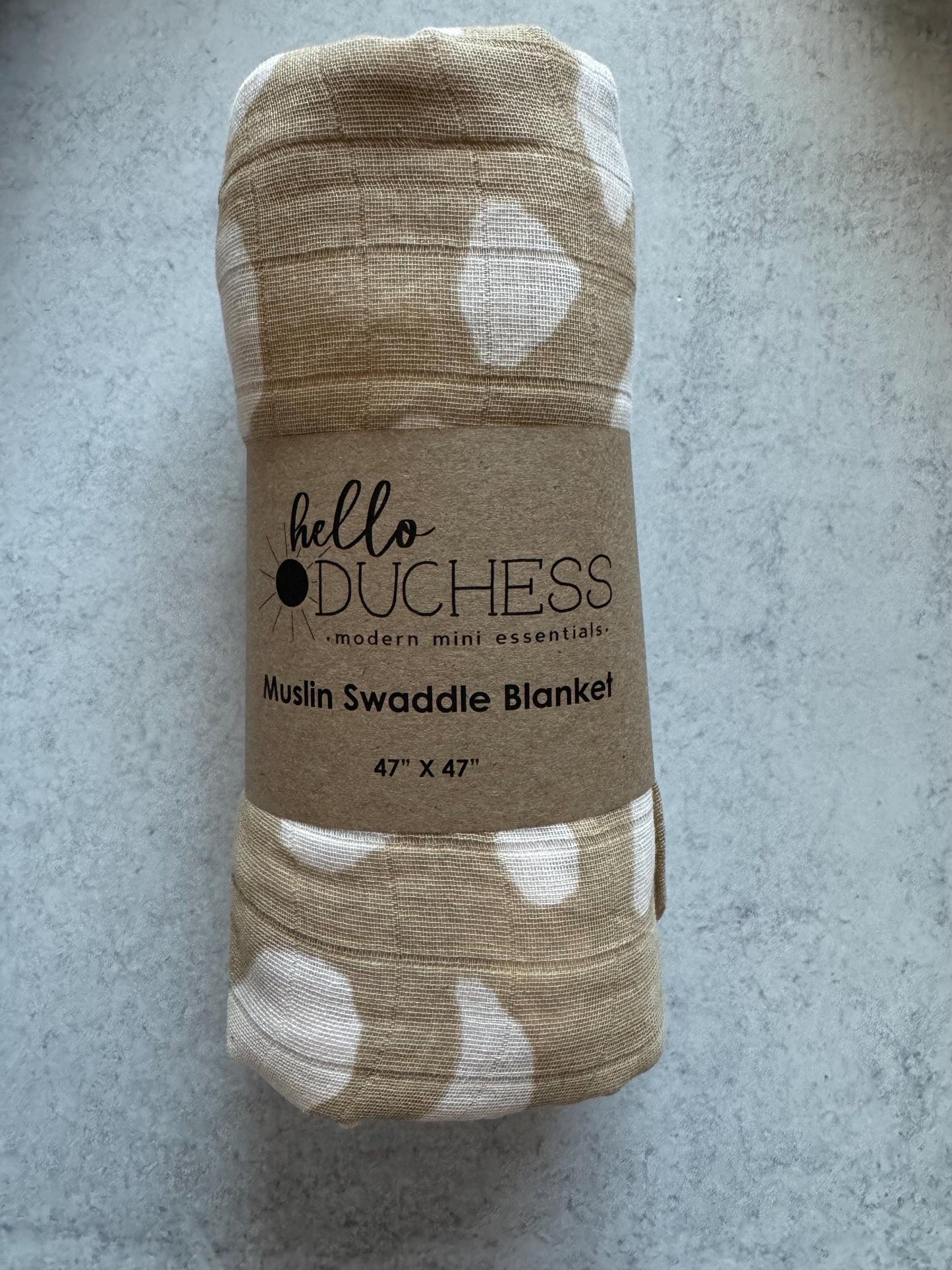 A beige and white animal print cotton swaddle blanket with the brand 'hello duchess' wrapped around it, placed on a grey background.
